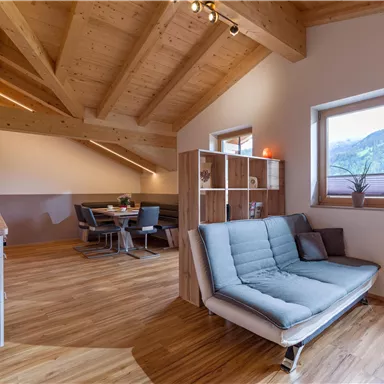 Ein modernes, helles Wohnzimmer mit Holzbalkendecke und gemütlichem Sofa. Im Hintergrund befindet sich ein Essbereich mit einem Tisch und Stühlen sowie großen Fenstern, die einen Blick auf die Berge bieten.