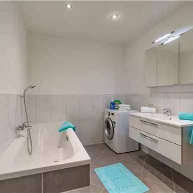 A modern bathroom with a bathtub and a white sink. There are towels in turquoise tones and a washing machine in the room.