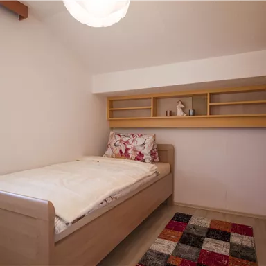 A simple bedroom with a single bed and a colorful carpet. The walls are light, and there are shelves above the bed.
