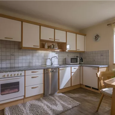 A modern kitchen with white cabinets and wood details. It is bright and inviting, with a small dining table and windows that let in plenty of light.