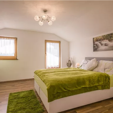 A modern bedroom with a large bed and green bedding. The furnishings include a desk and a cozy atmosphere.