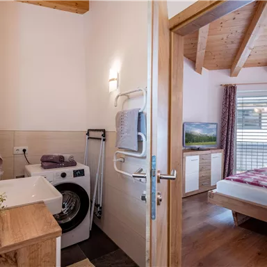 Ein modernes Badezimmer mit einer Waschmaschine und Holzverkleidung. Im Hintergrund ist ein Schlafzimmer mit einem Fernseher und einem Fenster zu sehen.