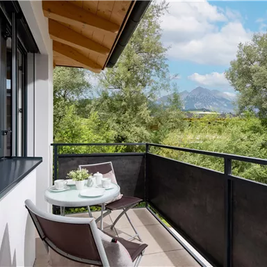 Ein gemütlicher Balkon mit einem kleinen Tisch und Stühlen, umgeben von grünen Bäumen. Im Hintergrund sind sanfte Berge und ein blauer Himmel zu sehen.