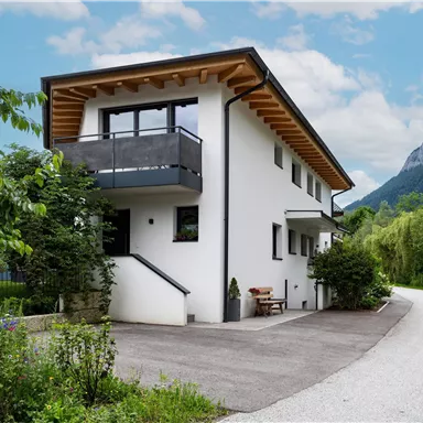 Ein modernes Haus mit Holzveranda und Garten, umgeben von Bäumen und Bergen. Die Zufahrtsstraße führt auf das Grundstück.