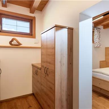 A cozy room with wooden cabinets and a window. In the background, a bedroom with a bed and gentle light sources can be seen.