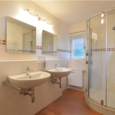 A modern bathroom with two sinks and large mirrors. There is a shower in the corner and bright, neutral colors on the walls.