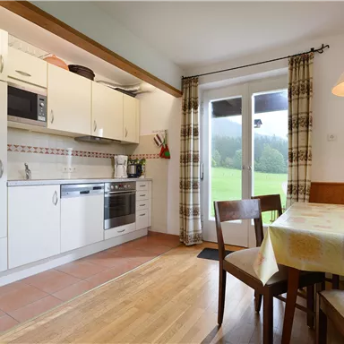 A modern kitchen with white cabinets and a wooden countertop. The dining area is furnished with a table and chairs, and large windows let in plenty of light.