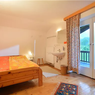 A cozy bedroom with a wooden bed and colorful bedding. Large windows offer a view of the greenery.