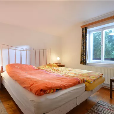A cozy bedroom with a double bed and colorful bedding. The room has a window with curtains and a small bedside table.