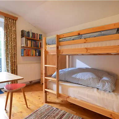 A cozy bedroom with a bunk bed and a small table. The room has large windows and a bookshelf.
