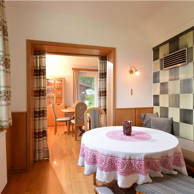 A cozy room with a round table and an elegant tablecloth. In the background, chairs and a decorative wall are visible.