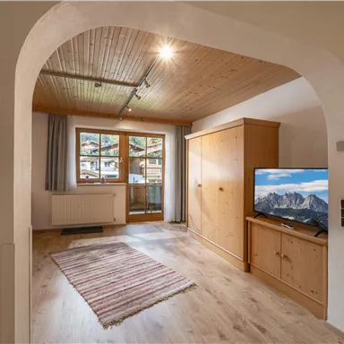 Ein gemütlicher Wohnraum mit Holzdecke und einem großen Fenster. Ein Fernseher und ein Holzschrank sind ebenfalls im Raum zu sehen.