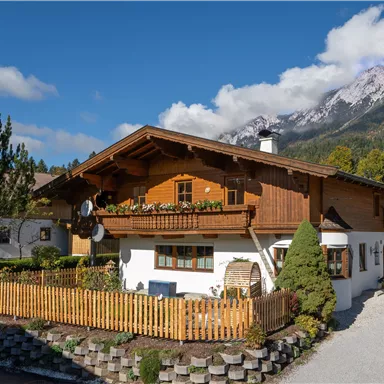 Ein traditionelles österreichisches Haus mit Holzbalkon und schönem Blumenschmuck. Im Hintergrund sind majestätische Berge unter einem blauen Himmel zu sehen.