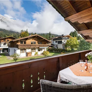 Ein gemütlicher Balkon mit einem Tisch und zwei Gläsern, umgeben von einer malerischen Berglandschaft. Im Hintergrund sind traditionelle Holzhäuser und üppiges Grün zu sehen.