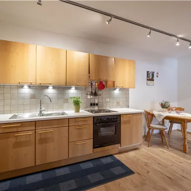 Eine moderne Küche mit Holzmöbeln und weißer Arbeitsplatte. Es gibt einen Esstisch und einen Teppich am Boden.
