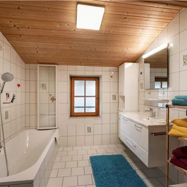 Ein modernes Badezimmer mit einer Badewanne und einer Dusche. Es gibt helle Fliesen, Holzdecke und farbige Handtücher.