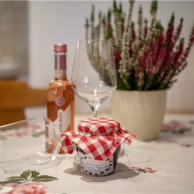 Ein gemütlicher Tisch mit einem Glas und einer Flasche Roséwein. Im Mittelpunkt steht ein Glas mit einem karierten Deckel und eine blühende Pflanze.