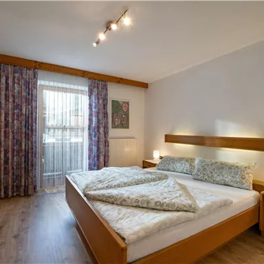 Ein gemütliches Schlafzimmer mit einem Doppelbett und Holzmöbeln. Große Fenster mit Vorhängen sorgen für viel Tageslicht.
