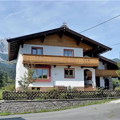 A cozy house in the mountains with wooden balustrades and a large garden. In the background, high mountains and a blue sky can be seen.