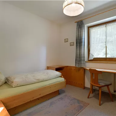 A simple bedroom with a single bed and a desk. The room has a large window with curtains and is designed in light wood tones.
