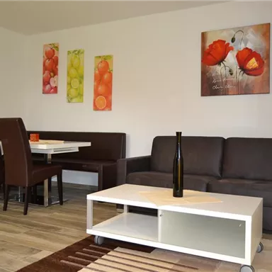 A cozy living room with a brown sofa and a modern coffee table. Colorful pictures of fruits and flowers hang on the wall.