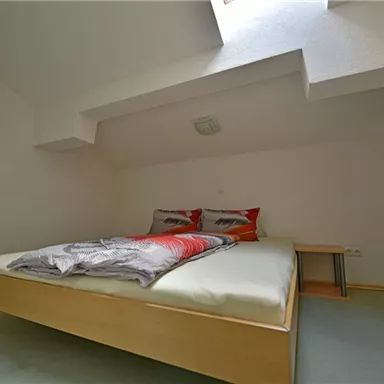 A simple bedroom with a double bed and colorful cushions. Next to the bed is a small table and the wall has a shelf.