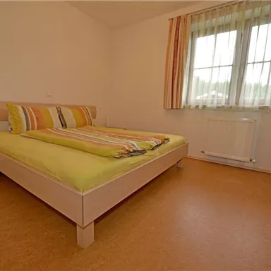 A bright bedroom with a large bed and colorful cushions. There is a bedside table and large windows with curtains.