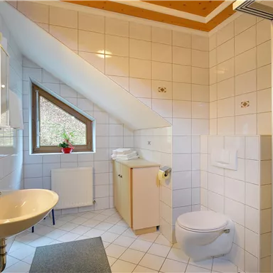 A modern bathroom with white tiles and a sloping ceiling. It features a sink, a toilet, and a window with a view outside.