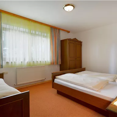 A simple bedroom with two beds and a wardrobe. The windows are decorated with light curtains and the floor is carpeted.