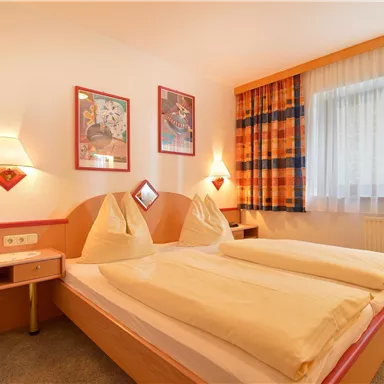 A cozy bedroom with two beds and colorful pillows. The walls are decorated with artwork, and the window lets in plenty of daylight.