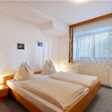 A bright bedroom with two beds and cozy bedding. The room has large windows with curtains and a warm atmosphere.