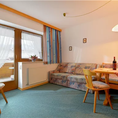 A cozy living space with a sofa, a table, and chairs. Large windows provide daylight and an inviting atmosphere.