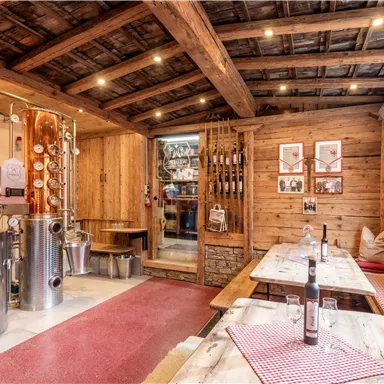 Ein gemütlicher Raum mit rustikalem Dekor und Holzwänden. Eine Brennerei steht im Vordergrund, während Tische mit roten Tischtüchern zum Sitzen einladen.