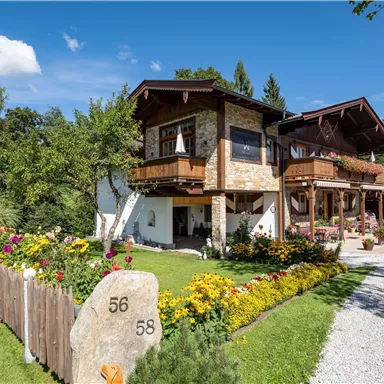 Ein schönes Haus mit Holzverkleidung und Balkon, umgeben von bunten Blumen. Der Himmel ist klar und blau, was eine entspannte Atmosphäre schafft.