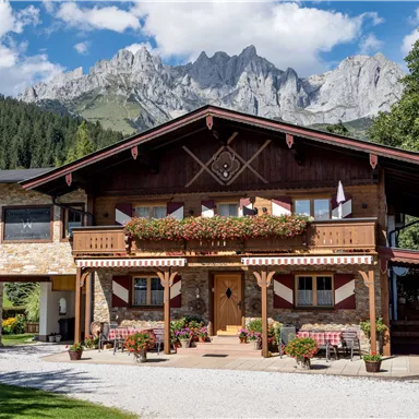 Ein charmantes Holzhaus mit schönen Blumenbalkonen. Im Hintergrund sind beeindruckende Berge und ein blauer Himmel zu sehen.