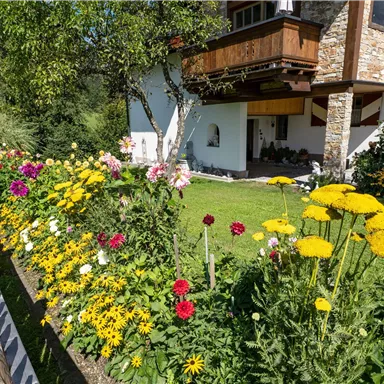 Ein blühender Garten mit bunten Blumen und einem gepflegten Grundstück. Im Hintergrund ist ein traditionelles Haus mit Balkon zu sehen.