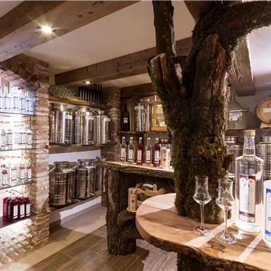 Eine gemütliche Brennerei mit Holzregalen und silbernen Behältern. Auf einem Tisch stehen Gläser und Flaschen von Spirituosen, umgeben von einem Baumstamm.