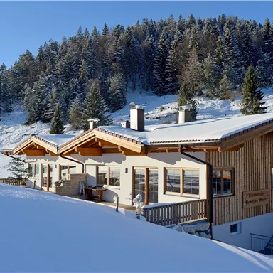 A cozy wooden house in a snow-covered landscape. Snow-covered trees are visible in the background.