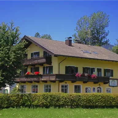 Ein sonniges Gebäude mit gelber Fassade und Balkonen, umgeben von Bäumen. An der Fassade sind Blumen Kästen mit bunten Pflanzen angebracht.
