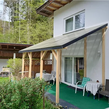Eine überdachte Terrasse mit weißen Stühlen und Tischen. Der Hintergrund zeigt eine weiße Wand und eine Doppel-Glastür.