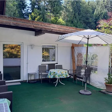 Eine gemütliche Terrasse mit Gartenmöbeln und einem Sonnenschirm sorgen für eine einladende Atmosphäre. Im Hintergrund sind grüne Bäume und eine Japanische Kirsche zu sehen.