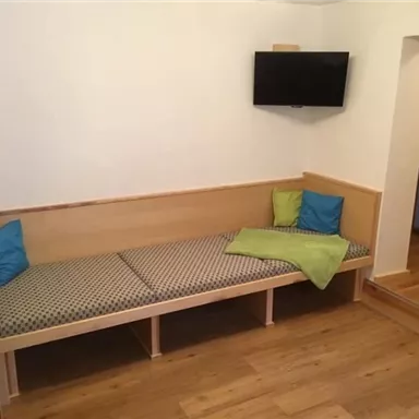 A simple living room with a bench and colorful cushions. A television hangs on the wall, and the floor is made of wood.