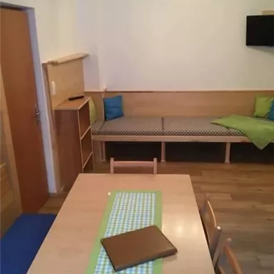 A simple dining room with a wooden table and chairs. There is a seating area and a television on the wall.