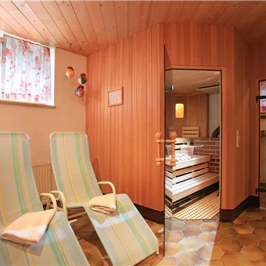 A cozy wellness area with loungers and a sauna in the background. The walls are made of wood, and there are bright windows with curtains.