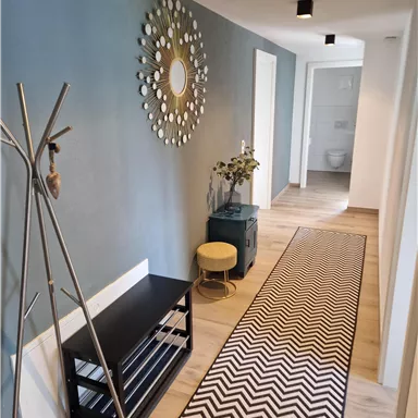 Ein moderner Flur mit einer grauen Wand und einem stilvollen Spiegel. Auf dem Boden liegt ein Teppich mit chevron-Muster und es stehen einige Möbelstücke, darunter eine Garderobe und ein kleiner Tisch.