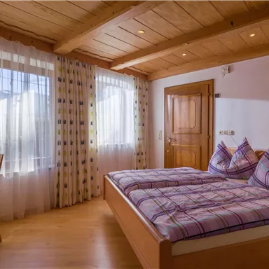 Ein gemütliches Schlafzimmer mit einem Doppelbett und zwei Kissen in lila Kariert. Große Fenster lassen viel Licht herein und ein Holzstuhl ergänzt die Einrichtung.