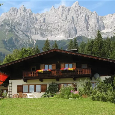 A traditional house in the mountains, surrounded by trees and meadows. In the background, majestic mountains rise under a clear sky.