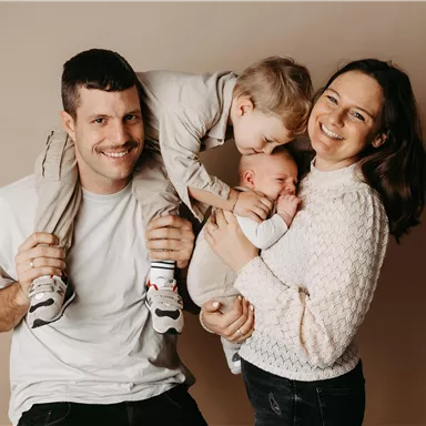 Eine fröhliche Familie posiert zusammen. Der Vater trägt ein Kind auf den Schultern, während die Mutter das Baby in ihren Armen hält.