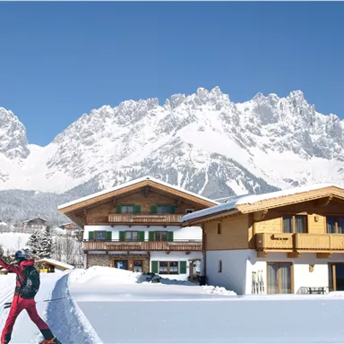 A snowy winter landscape with majestic mountains in the background. Two people in ski attire enjoy the view in front of an alpine chalet.