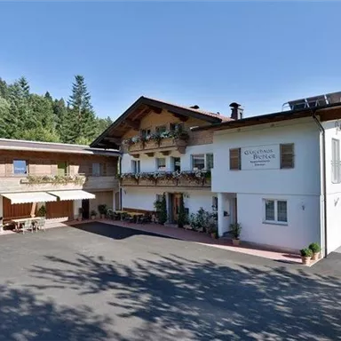 A charming building in alpine style with balconies and an inviting façade. Surrounded by trees and mountains in clear blue sky.